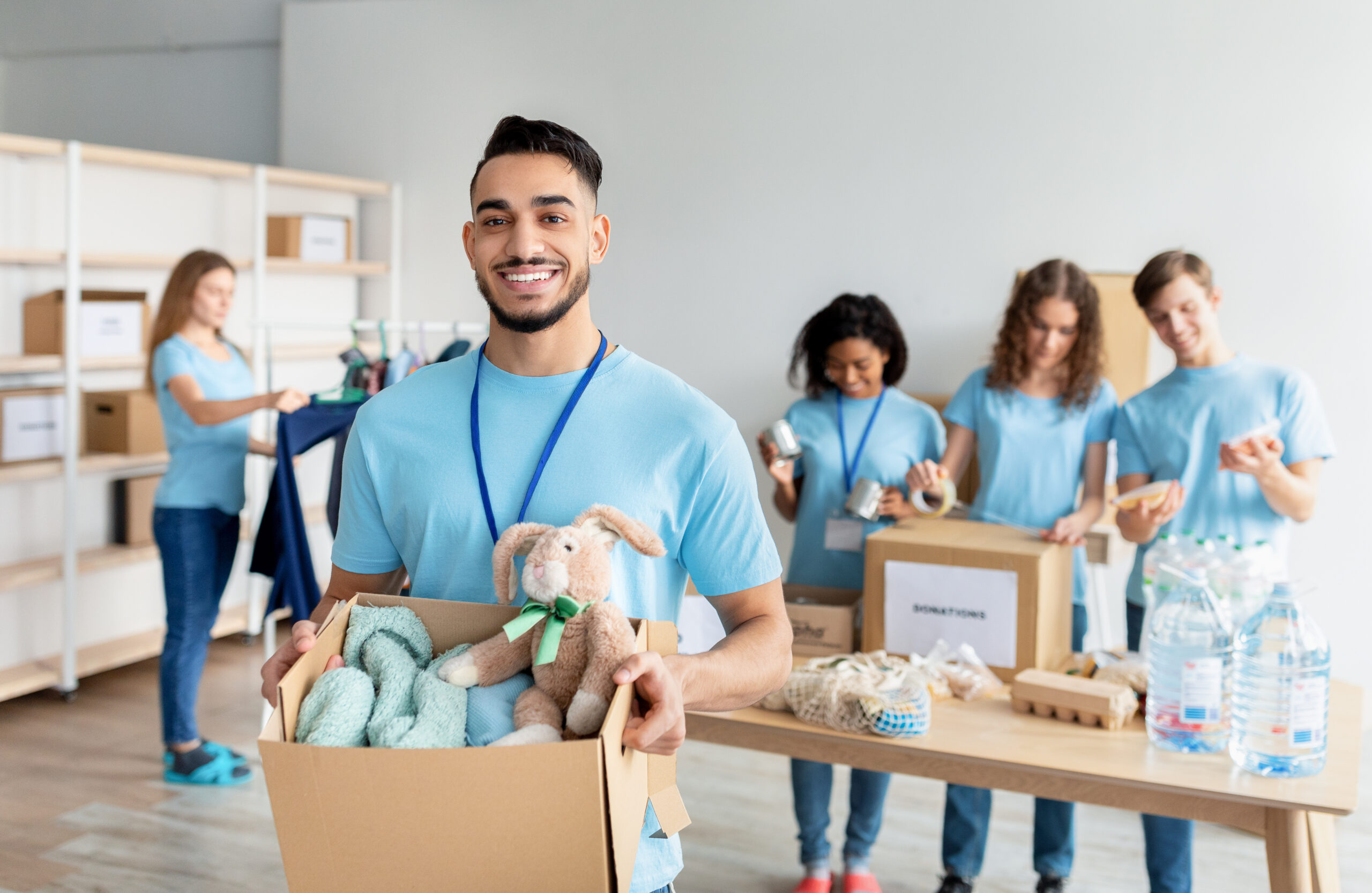 Arab male volunteer holding box with clothes and toys for donation, smiling to camera while working at charity center with group of colleagues, packing products in boxes on background